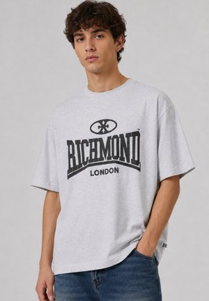 Giovane uomo con capelli ricci che indossa una T-shirt oversize grigio chiaro con testo e logo "Richmond London" in nero, abbinata a jeans blu.