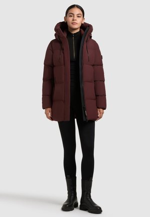 Chaqueta acolchada burdeos con capucha, que presenta un diseño acolchado, cremallera frontal y detalles en negro. Lleva puesta sobre un conjunto negro y combinado con botas negras.