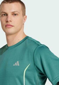 T-shirt de sport à manches courtes en turquoise, fabriqué en tissu lisse. Il présente des panneaux d'épaule texturés et un logo discret sur la poitrine.