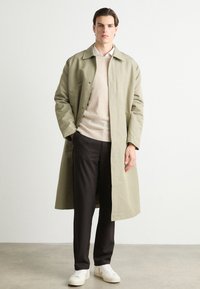 Licht olijfgroene trenchcoat met een rechte snit, knoopsluiting en zijzakken, gedragen over een beige trui en zwarte broek. Witte sneakers.
