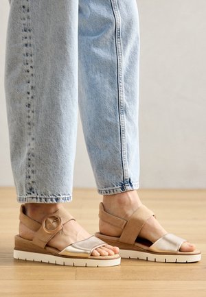 Sandali marroni con una cintura in tessuto lucido, una banda tessile ampia e una fibbia circolare su una suola piatta con dettagli testurizzati.