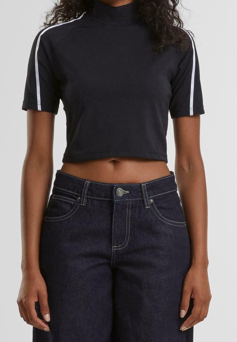 T-shirt noir court avec col haut, manches courtes et rayures blanches sur les épaules, associé à un jean en denim foncé avec des coutures claires.