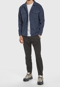 Marinblå skjorta med knappar och två bröstfickor, bärs över en vit t-shirt och matchas med svarta smala byxor och grå sneakers med blå detaljer.