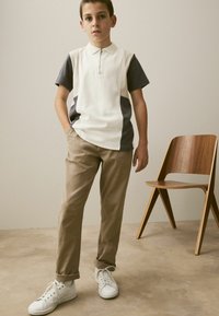 Polo shirt à col avec une fermeture éclair, corps blanc et manches gris foncé, associé à un pantalon beige et des baskets blanches. Chaise en bois en arrière-plan.
