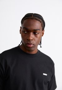 Zwarte katoenen t-shirt met een ronde hals en korte mouwen, voorzien van een wit "OBEY" logo op de linkerkant van de borst. Soepele textuur.