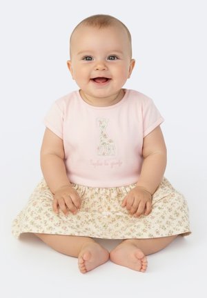 Bambino sorridente seduto con le gambe incrociate, indossa un top rosa con disegno di giraffa e una gonna con motivo floreale.