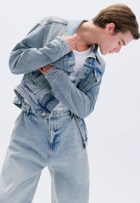 Giacca in denim celeste chiaro indossata sopra una t-shirt bianca, abbinata a jeans a vita alta dello stesso colore con dettagli strappati.