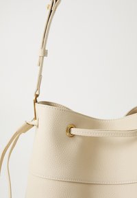 Bežová kabelka v tvare vedra vyrobená z textúrovanej kože, so zlatými kovovými doplnkami a nastaviteľným popruhom. Hladký, nezdobený povrch.