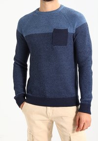 Pull en maille bleu marine avec une finition texturée, comprenant une poche patch rectangulaire bleu clair et des détails en côtes aux poignets et à l'ourlet.