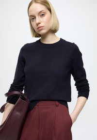 Tengerészkék pulóver puha kötött anyagból, kerek nyakkivágással és hosszú ujjakkal, bordó, szabott nadrággal és texturált bordó táskával párosítva.