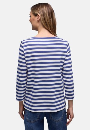 Žena se světle hnědými vlasy, oblečená v modrobílém pruhovaném tričku s dlouhým rukávem a modrých džínách, pohled zezadu.