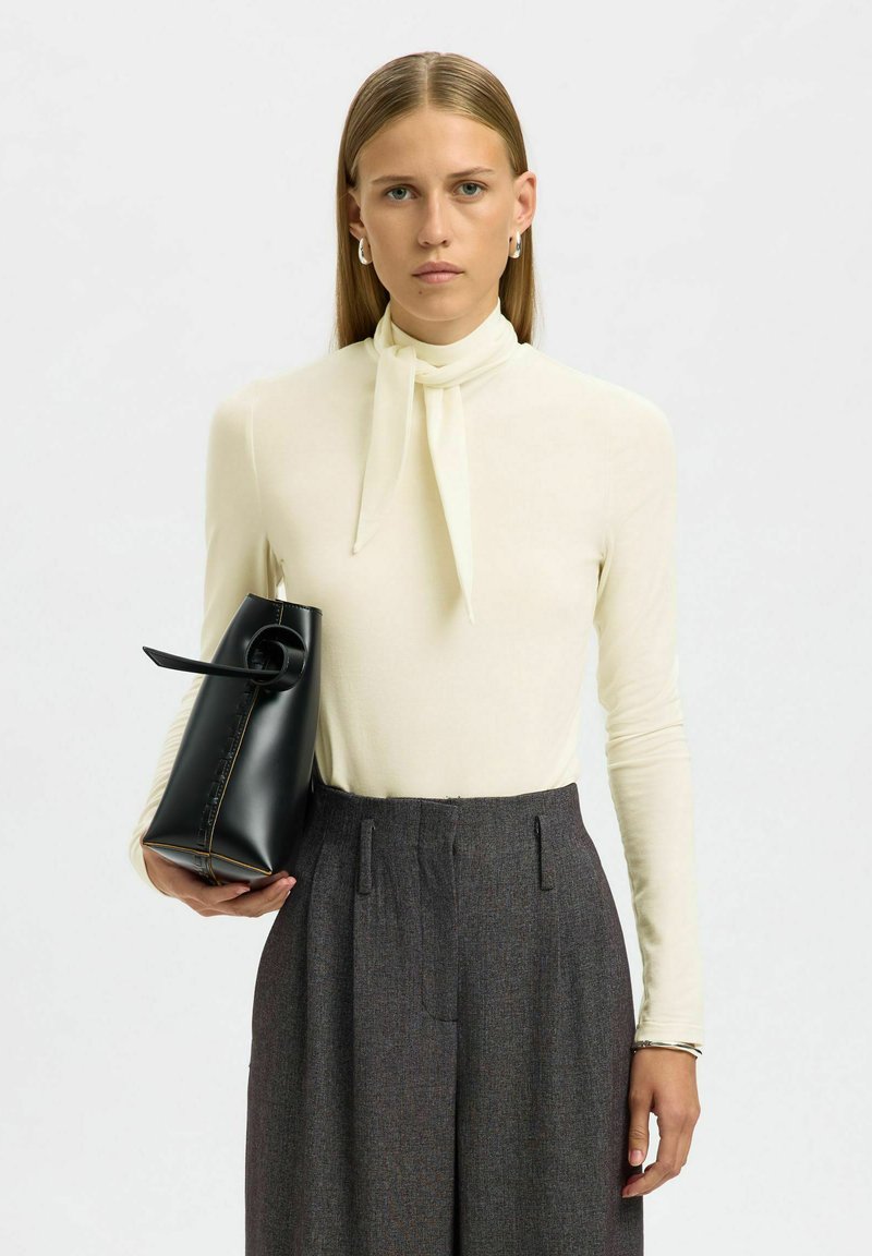 Haut crème à manches longues avec un col noué, pantalon gris ajusté et un sac à main noir avec une anse en boucle et des détails de couture jaune.