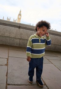 Kootud kampsun oliivrohelises, tumesinis ja valgetes triibudes; teksapüksid; sinised kingad Velcro kinnitustega; lapse seisab, taustal Big Ben.