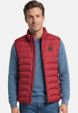 Mann trägt eine rote gesteppte Weste über einem blauen Strickpullover und blauen Jeans vor einem einfachen grauen Hintergrund.