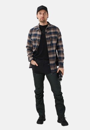 Man met zwarte pet, beige en navy geruit overhemd, zwarte broek, houdt zwarte waterfles vast, staand tegen een witte achtergrond.