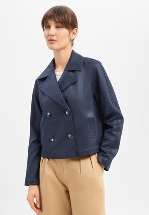 Kobieta z krótkimi brązowymi włosami, ubrana w granatową, dwurzędową marynarkę i beżowe spodnie, stojąca z jedną ręką w kieszeni na białym tle.