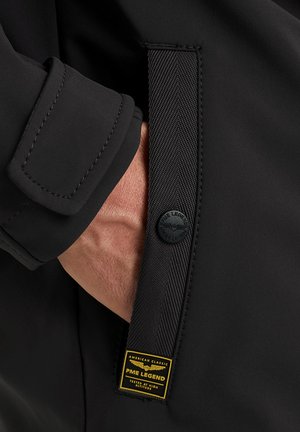 Hand in schwarzer Jackentasche mit strukturiertem Riemen mit Druckknopf und gelbem PME Legend Etikett mit der Aufschrift "American Classic Tested at High Altitude."