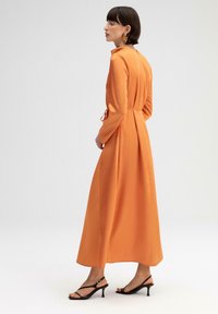 Lang oranje jurk met lange mouwen, knoop in de taille, gladde textuur en een zijsplit. Gepaard met zwarte sandaaltjes met banden.