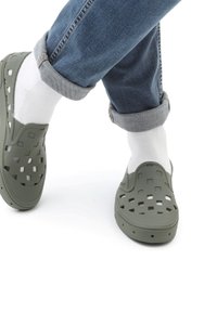 Olive green clogs with square cutouts, made of lightweight material, worn with white socks. Rolled cuff of blue jeans visible.