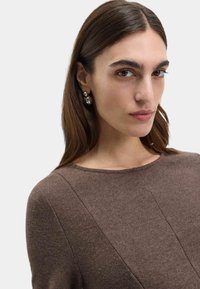 Brązowy sweter z okrągłym dekoltem, ozdobiony teksturowymi wzorami i ukośnymi szwami. Srebrne kolczyki dodają metalicznego akcentu.