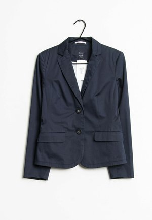 Blazer bleu marine pour femme avec deux boutons, revers crantés et poches à rabat à l'avant, suspendu sur un cintre noir devant un fond blanc.