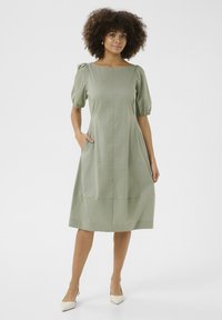 Olivno zelena obleka s kratkimi napihnjenimi rokavi, okroglim izrezom in stranskimi žepi. Gladek material s A-silhueto do kolen.