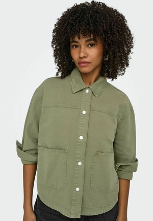 Młoda kobieta o kręconych włosach, ubrana w oliwkowo-zieloną kurtkę z guzikami, z podwiniętymi rękawami i dużymi przednimi kieszeniami, na neutralnym tle.