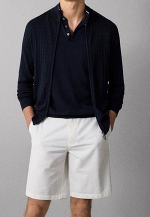 Mężczyzna ubrany w czarny sweter na zamek, pod którym ma czarną koszulkę polo, oraz białe szorty sięgające do kolan, stoi z rękami w kieszeniach.