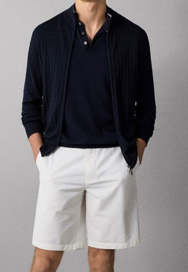 Homme portant un pull noir à fermeture éclair sur un polo noir et un short blanc arrivant aux genoux, debout les mains dans les poches.