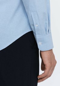 Lichtblauw overhemd met lange mouwen, gemaakt van katoen, met knoopsluitingen bij de manchetten en een gebogen zoom. De stof heeft een gestructureerd patroon.