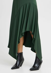 Mörkgrön asymmetrisk kjol med flödande tyg och sidodraperingar, i kombination med svart spetsig ankelboots med en kraftig klack.