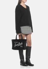 Pull noir en maille, jupe à carreaux, bottes noires hauteur genou et un sac fourre-tout noir avec texte blanc et bords effilochés.