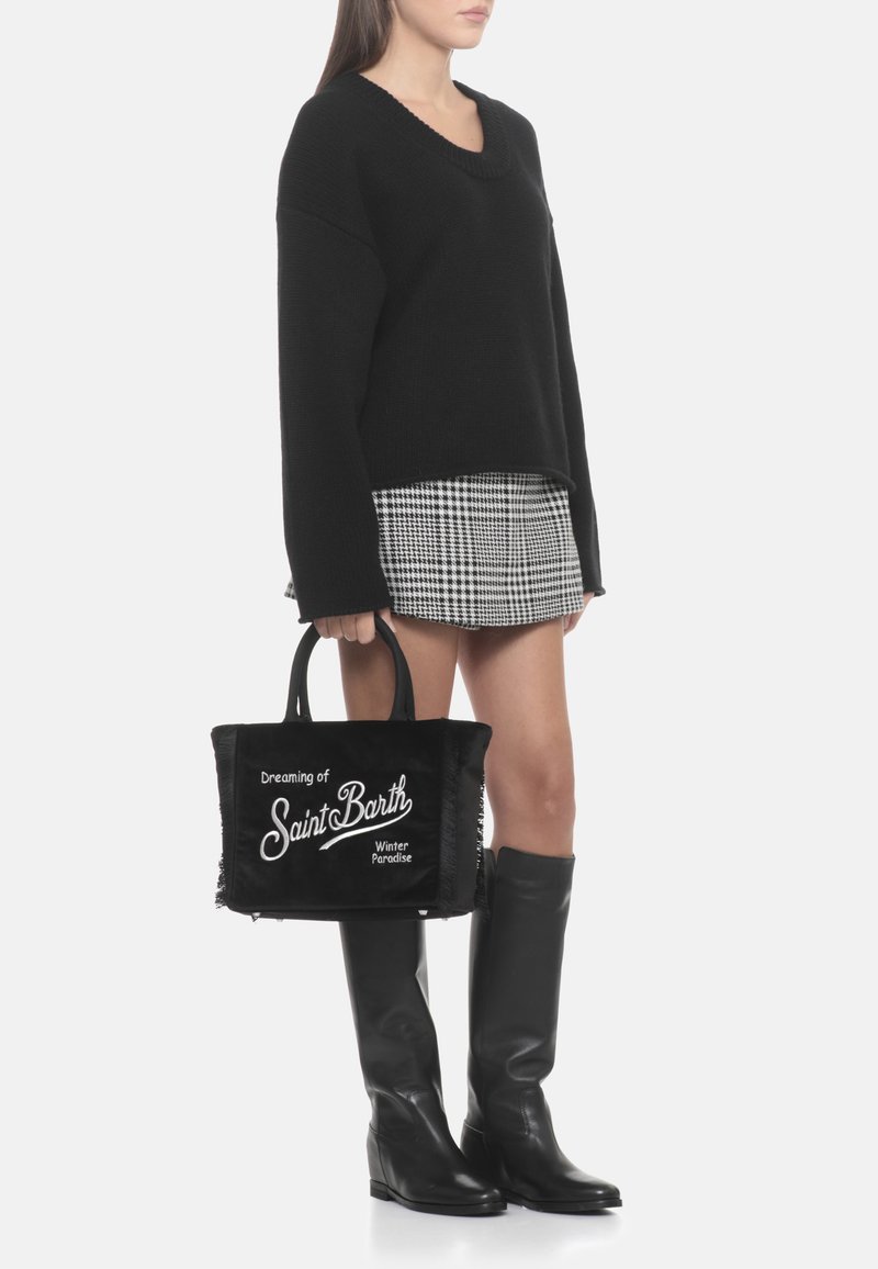 Pull noir en maille, jupe à carreaux, bottes noires hauteur genou et un sac fourre-tout noir avec texte blanc et bords effilochés.