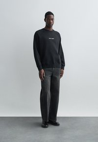 Μαύρο φούτερ με λευκό λογότυπο, μακριά μανίκια, ριμπ ύφανση στους αντίχειρες, συνδυασμένο με σκούρες γκρι φαρδιές παντελόνες και μαύρα παπούτσια, σε στάση απέναντι από ανοιχτό φόντο.