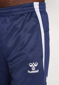 Jūriniai sportiniai šortai su baltais šonų juostelėmis, pagaminti iš lygaus audinio. Apatinėje kairėje dalyje yra baltas "hummel" logotipas.