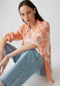 Blusa floreale arancione leggera e trasparente con maniche lunghe, abbinata a jeans dritti blu chiaro. Mettendo in risalto il motivo floreale e la texture del tessuto.