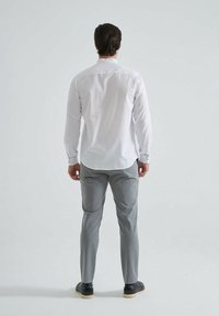 Chemise blanche à manches longues avec des poignets boutonnés, associée à un pantalon gris clair et des chaussures noires, vue de dos sur un fond uni.