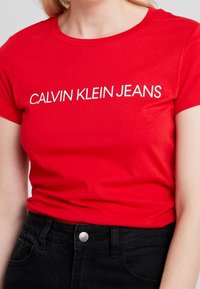 Red cotton t-shirt with short sleeves featuring white "CALVIN KLEIN JEANS" lettering across the chest, paired with black denim pants.
