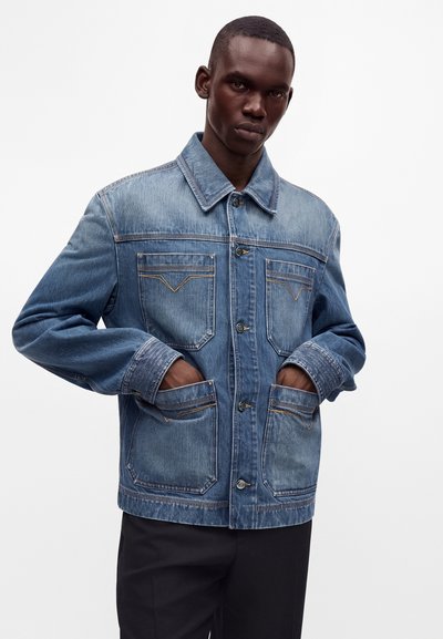Homme portant une veste en jean bleue avec quatre poches avant, debout les mains dans les poches, devant un fond blanc uni.