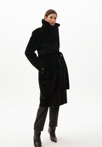 Černý umělý kožíšek s vysokým límcem, páskem v pase, náprsenkovými kapsami a zapínáním na knoflíky. Nosený přes černé vzorované kalhoty a kotníkové boty.