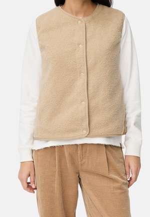 Personne portant un gilet en polaire beige boutonné sur une chemise blanche à manches longues et un pantalon en velours marron, debout devant un fond uni.