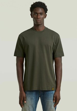 Jonge man met kort krullend haar, gekleed in een effen donkergroen T-shirt met korte mouwen en een blauwe spijkerbroek, staand tegen een effen achtergrond.