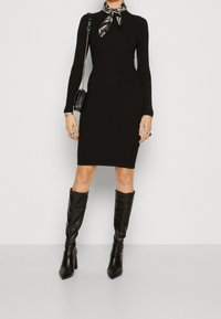 Vestito nero a coste con maniche lunghe e collo alto, abbinato a una sciarpa nera a fantasia e stivali neri alti fino al ginocchio con punta, completato da una piccola borsa.