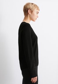 Svart genser med rund hals, ribbestrikkede ermer og nederkant. Materialet ser mykt ut, med et enkelt og rent design. Modellen har på seg svarte ribbestrikkede bukser.
