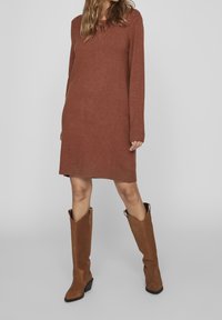 Abito in maglia marrone con maniche lunghe, polsini e orlo a coste, abbinato a stivali alti in suede marrone con un leggero tacco.