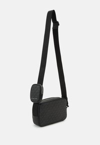 Fekete sling táska állítható pánttal, melyet struktúrált monogramos minta és sima bőrkiegészítők jellemeznek. Elülső zipzáras zsebet tartalmaz.