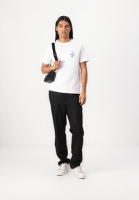 T-shirt en coton blanc avec un logo teal, accompagné d'un pantalon noir et de baskets blanches ; tenant un petit sac bandoulière noir.