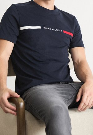 Mężczyzna ubrany w granatową koszulkę Tommy Hilfiger z białymi i czerwonymi paskami, siedzący na beżowym krześle, widoczne szare dżinsy.