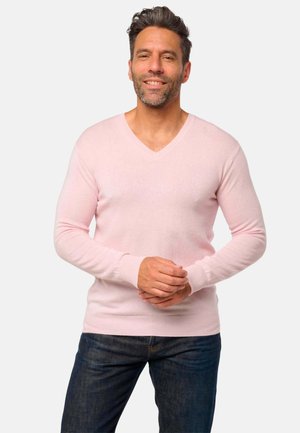 Homme souriant avec une barbe, portant un pull V en coton rose clair et un jean sombre, se tenant les mains jointes devant un fond clair.