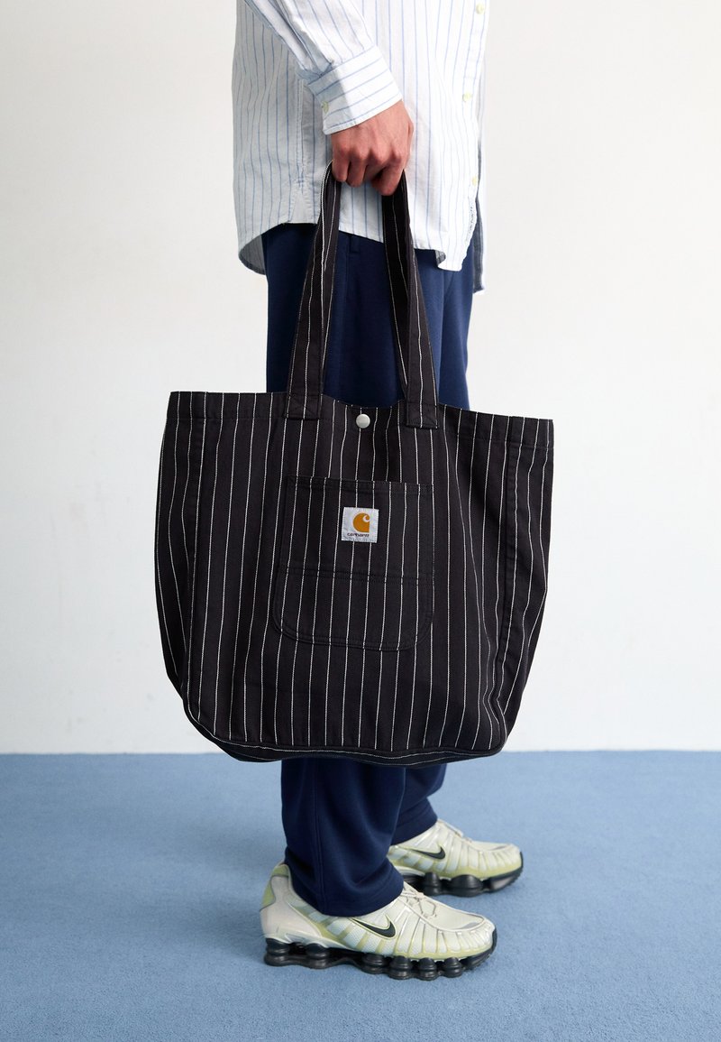 Sac fourre-tout à rayures noires avec une poche avant et un patch logo, fabriqué en tissu durable. Deux poignées robustes pour un transport facile.